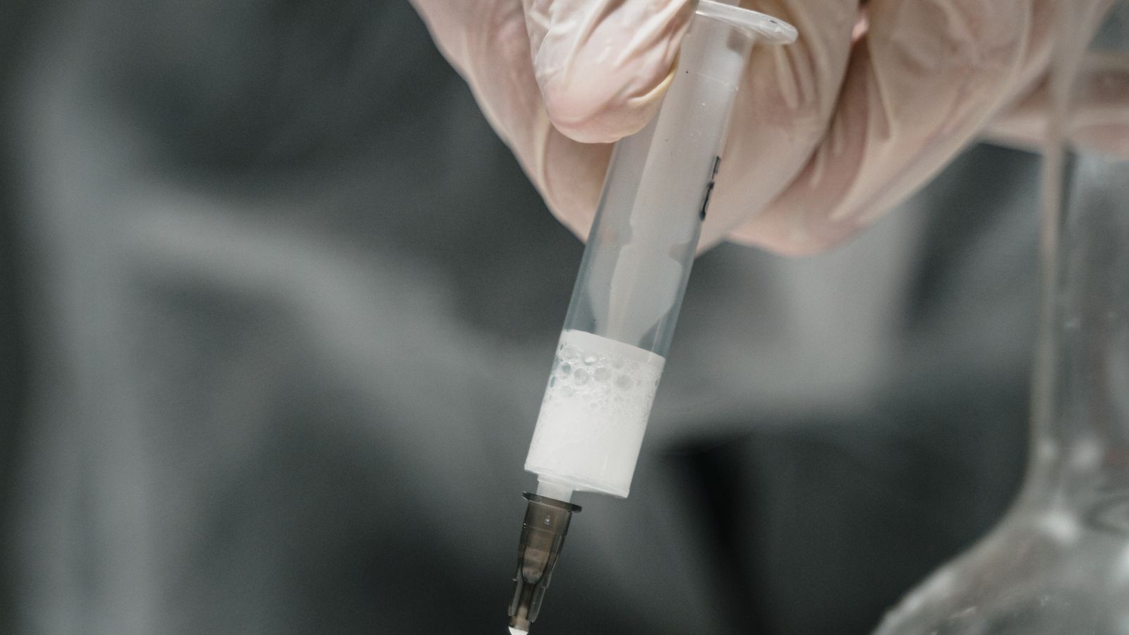 Close-up of gloved hands using a syringe to draw liquid from a small glass vial on a lab bench.